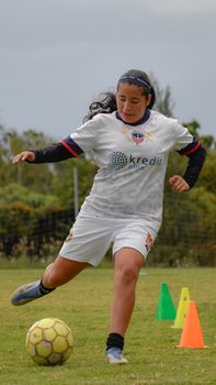 A young woman soccer player running drills on a grassy field beside colorful cones.