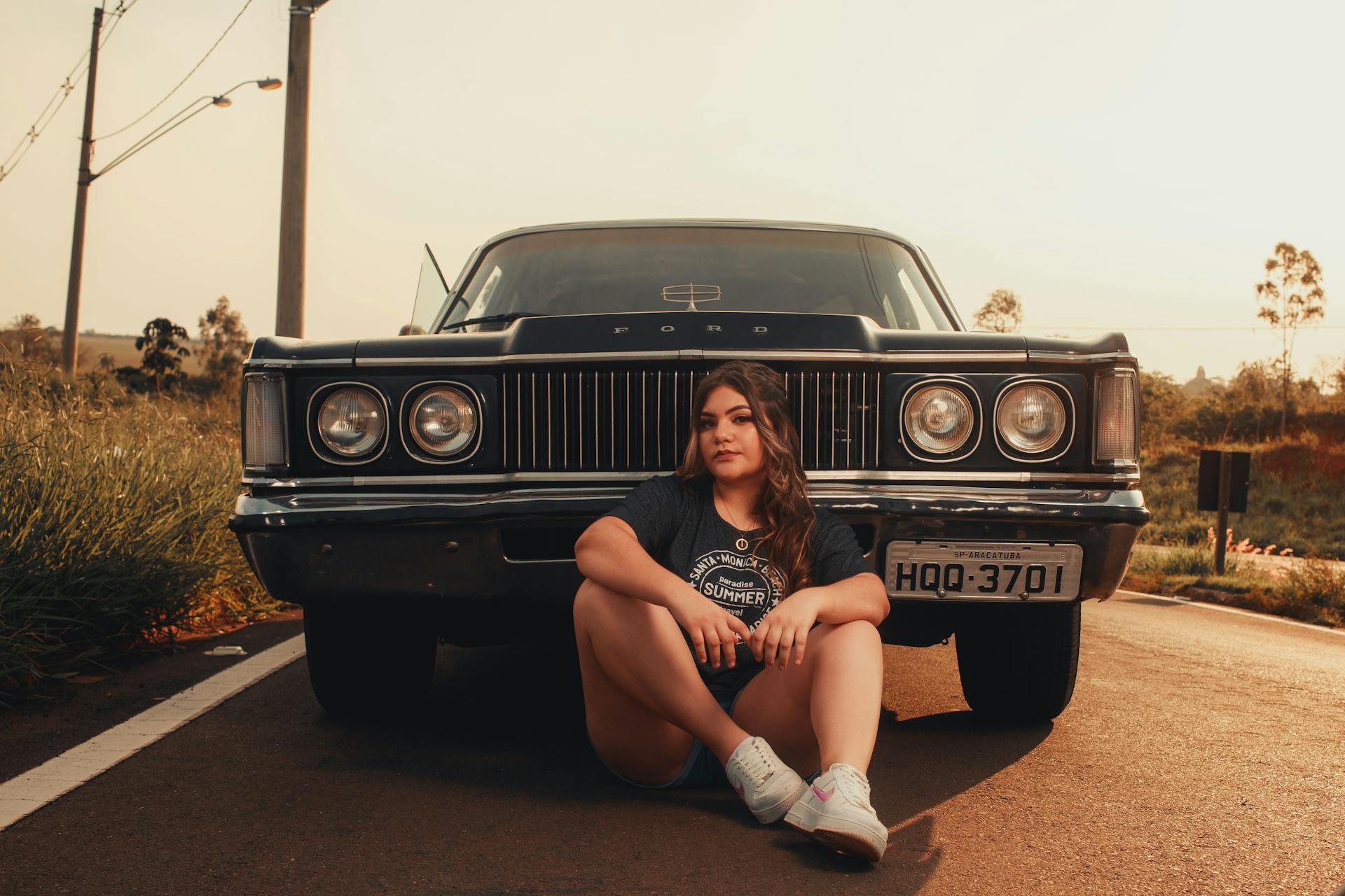 A woman sits in front of a classic Ford car on a rural road during a sunny day.