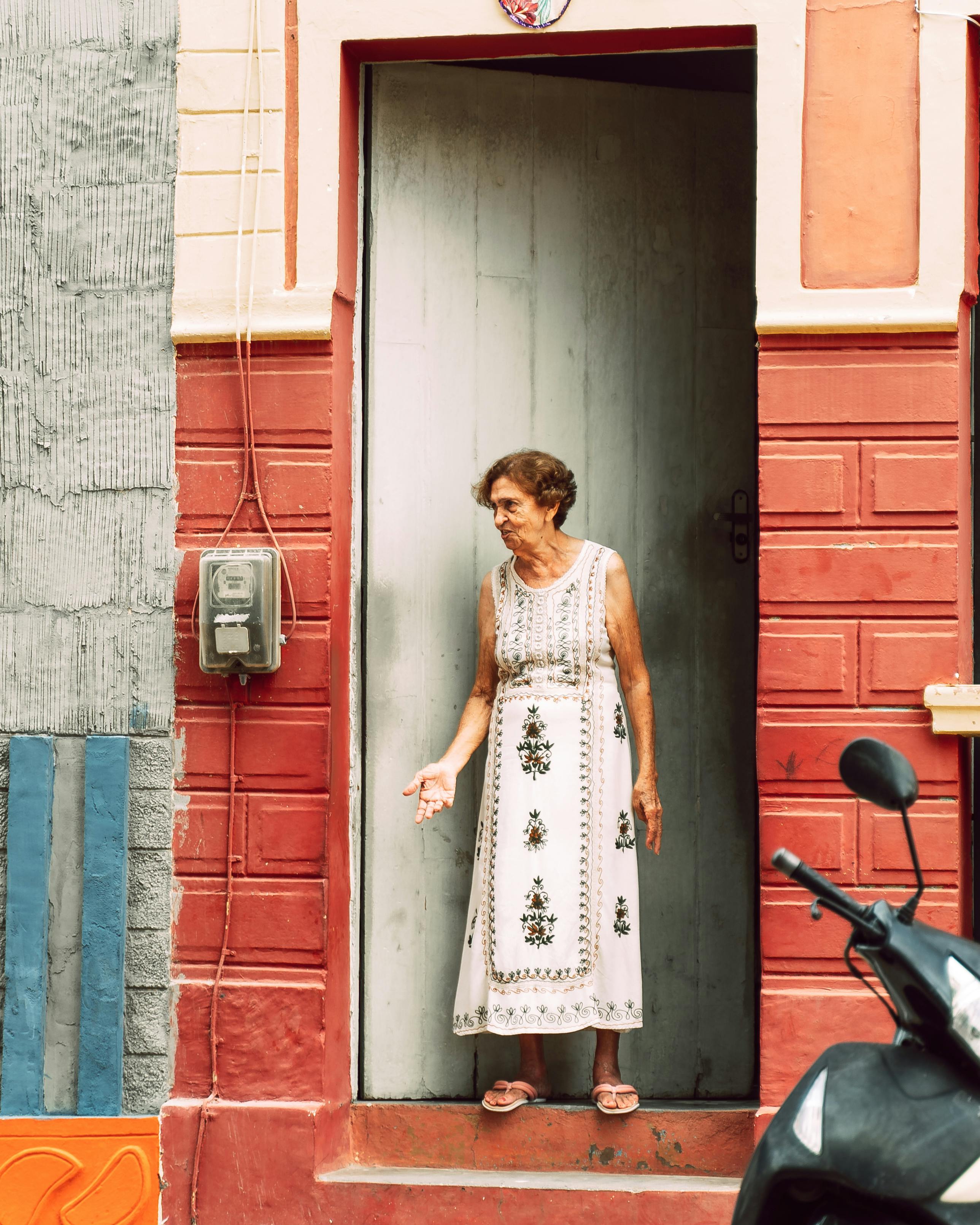 Elderly woman standing in a colorful doorway on a sunny day.