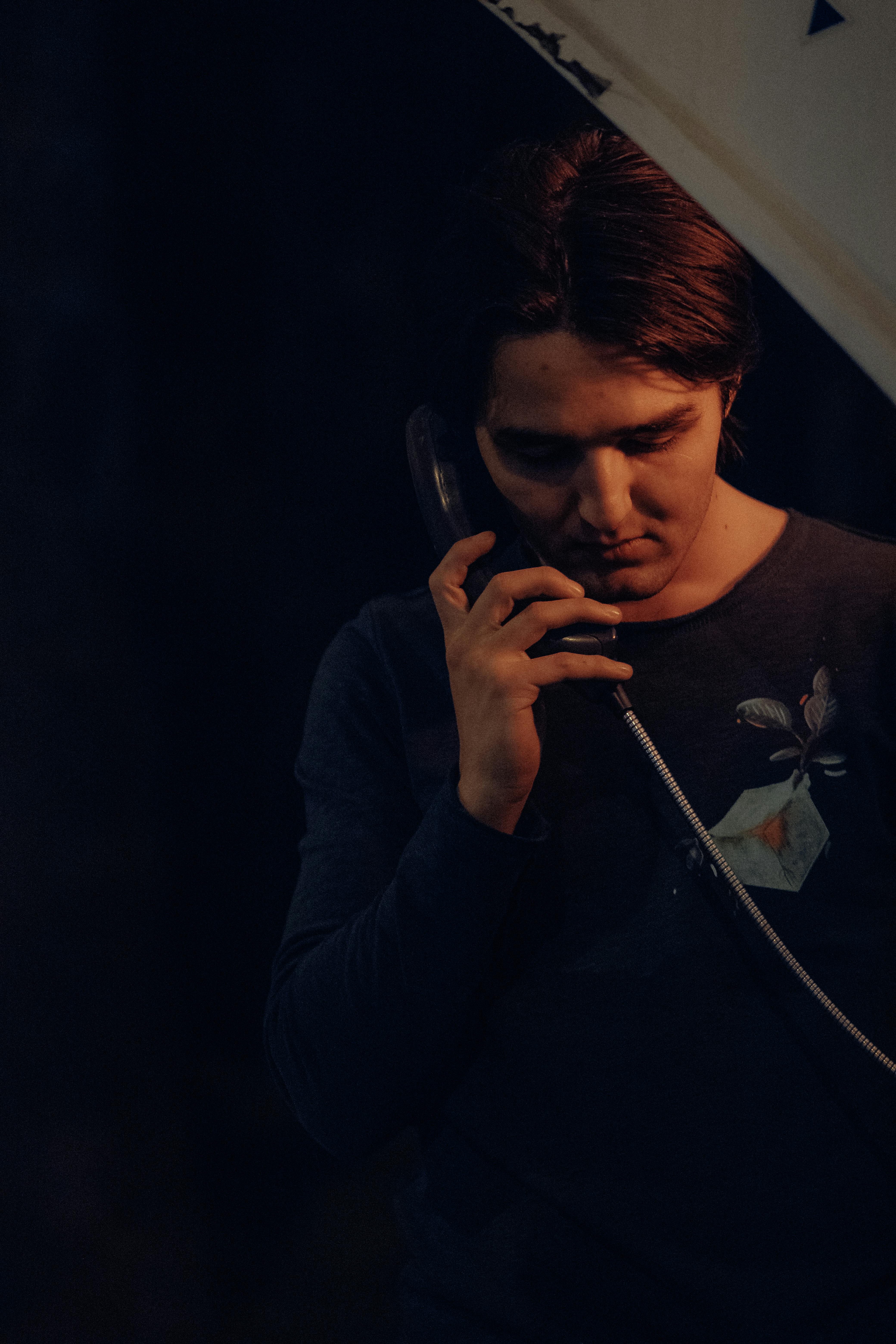 Free A young man stands in dim lighting holding a landline phone, creating a moody atmosphere. Stock Photo