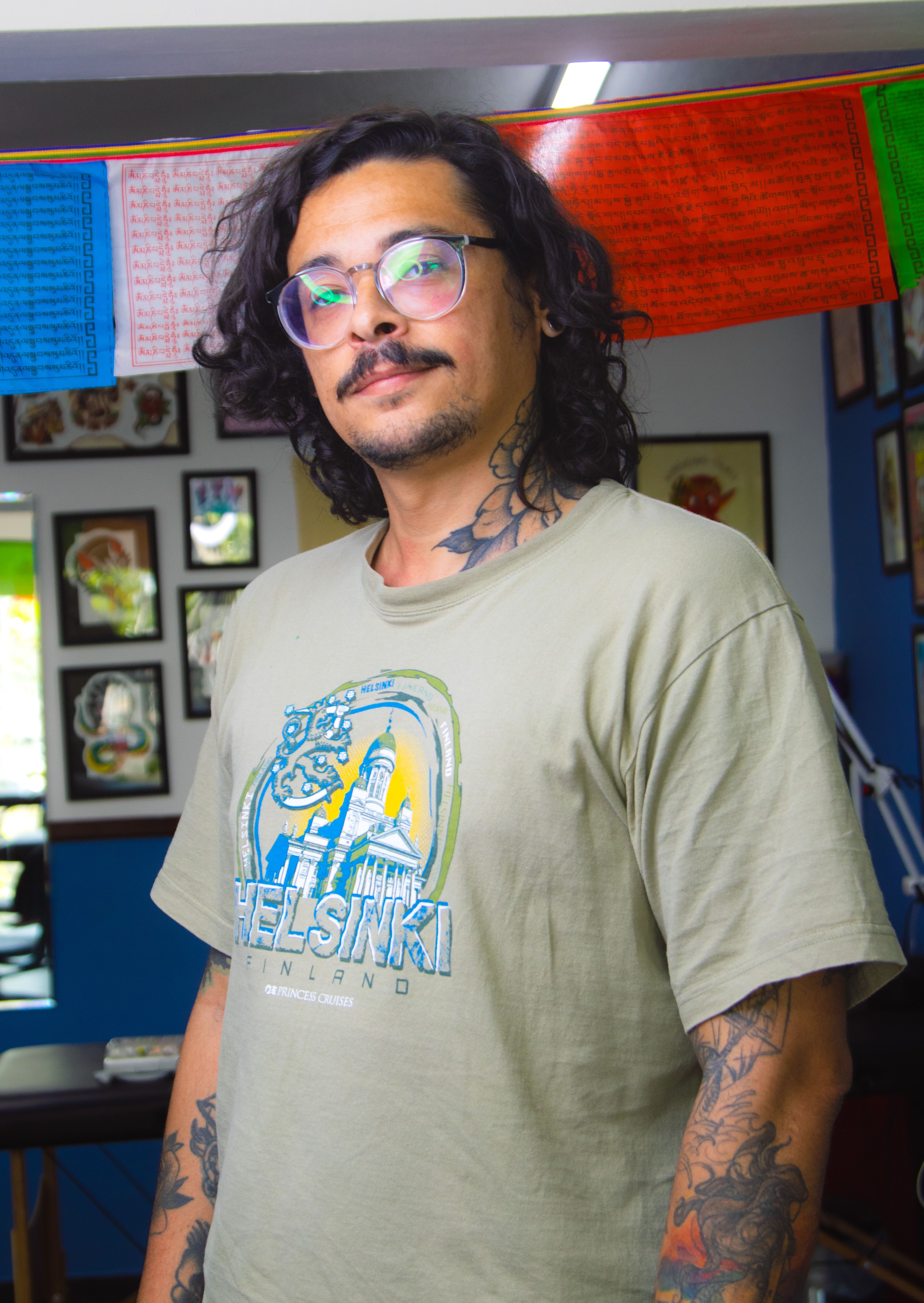 Young man with tattoos and eyeglasses standing indoors in casual wear.