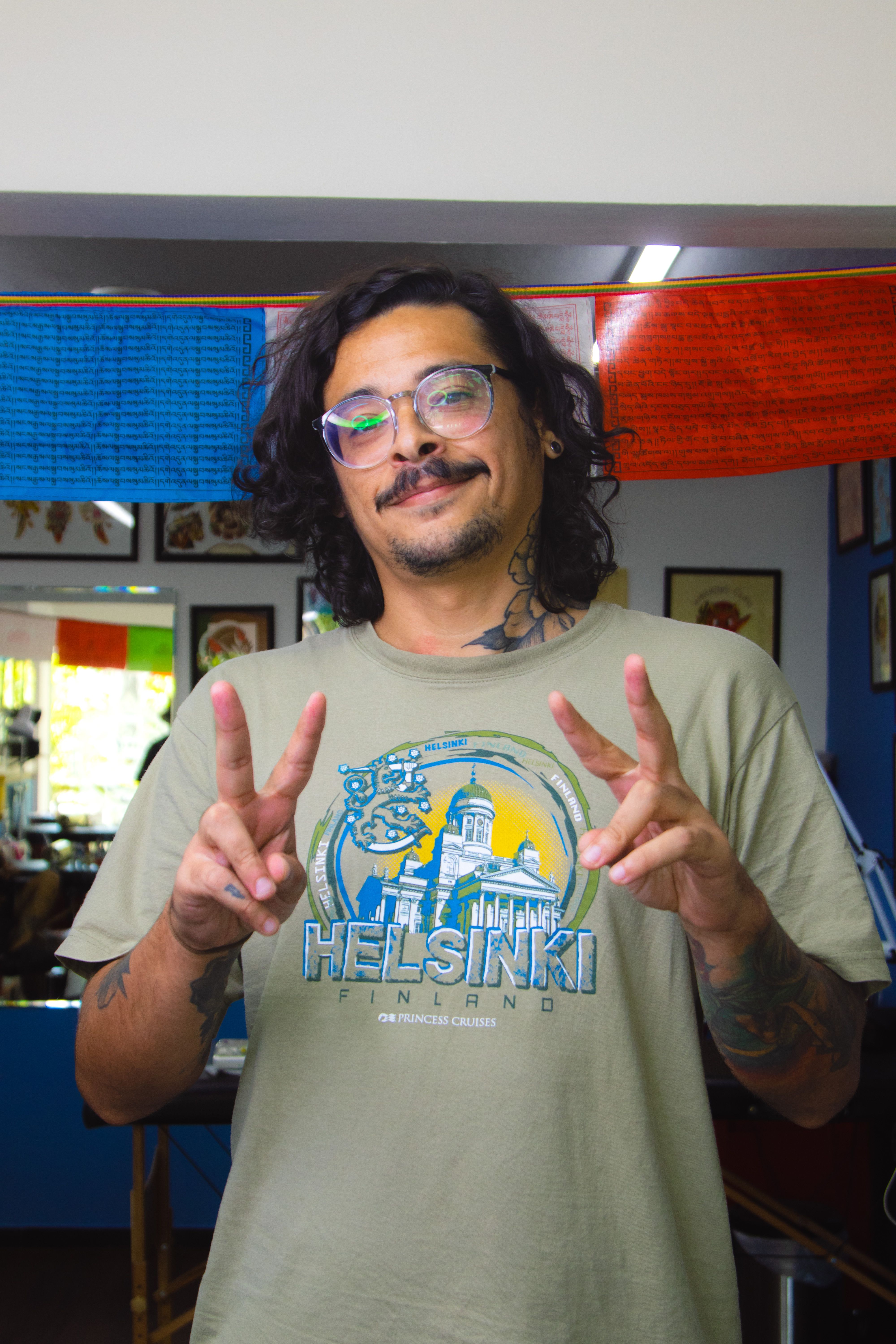 A smiling man with tattoos and glasses wearing a Helsinki t-shirt makes a peace sign indoors.