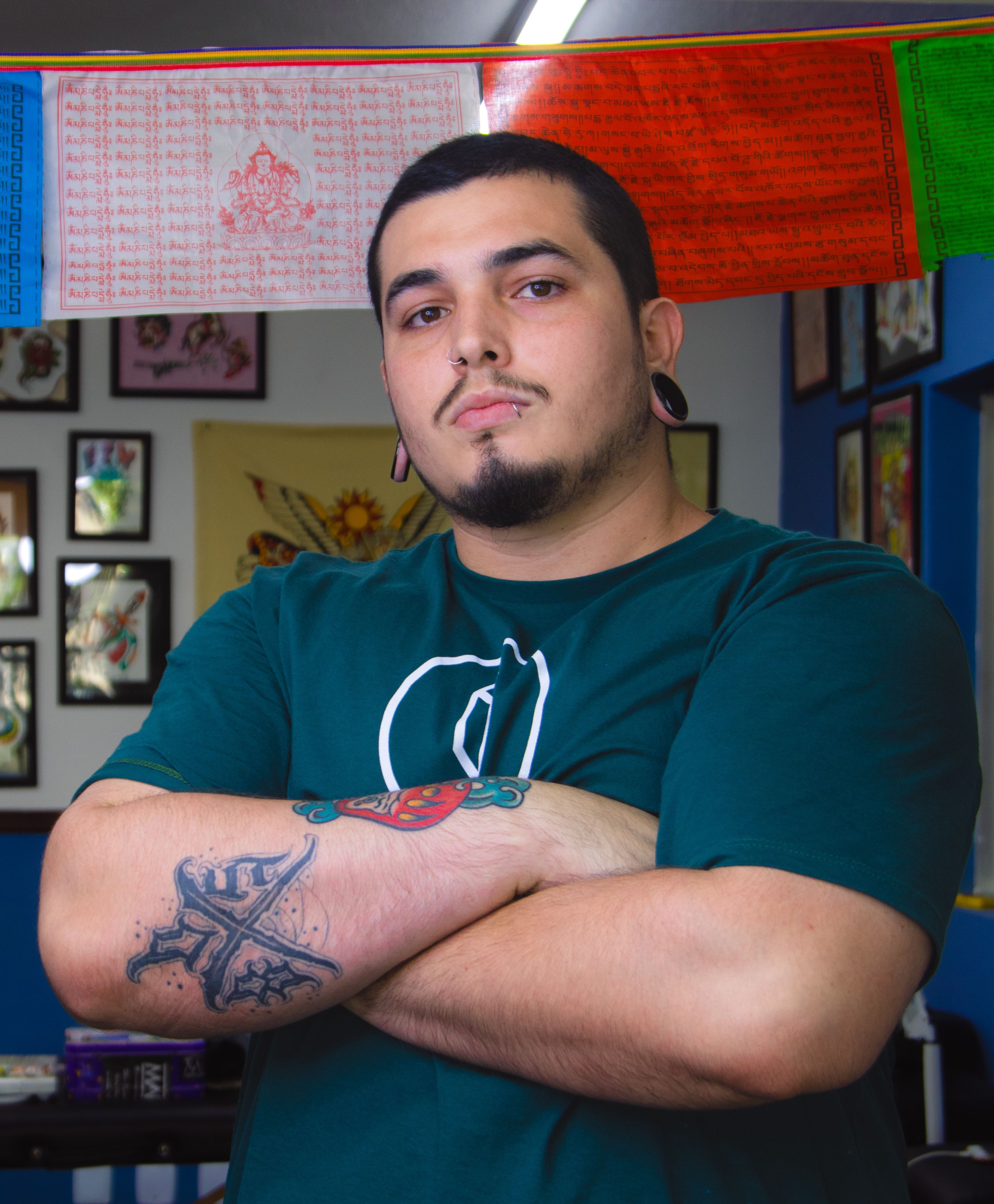 Tattooed man with folded arms standing confidently in an artistic studio with vibrant decor.