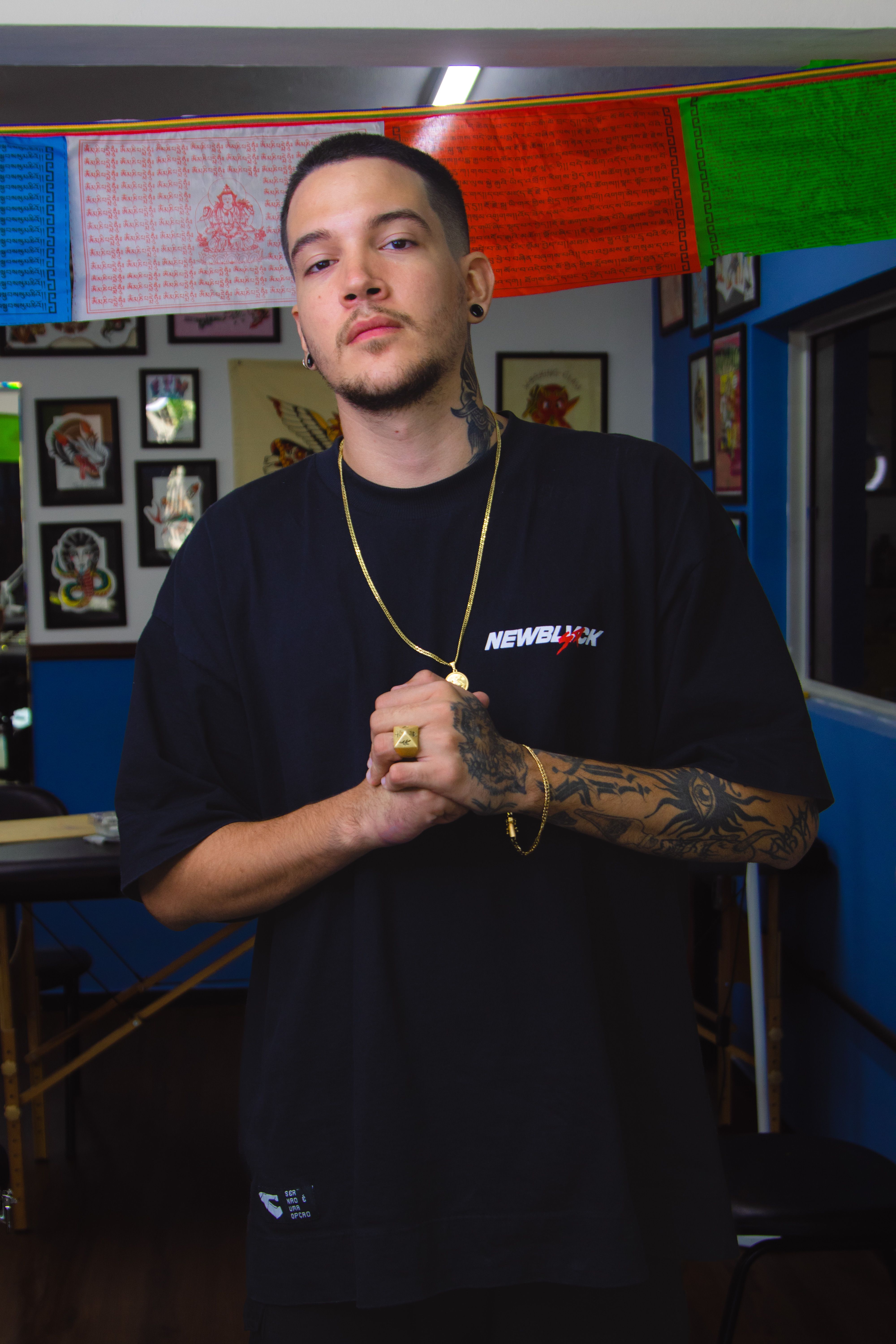 Man with tattoos standing confidently in a tattoo studio wearing a black t-shirt and accessories.