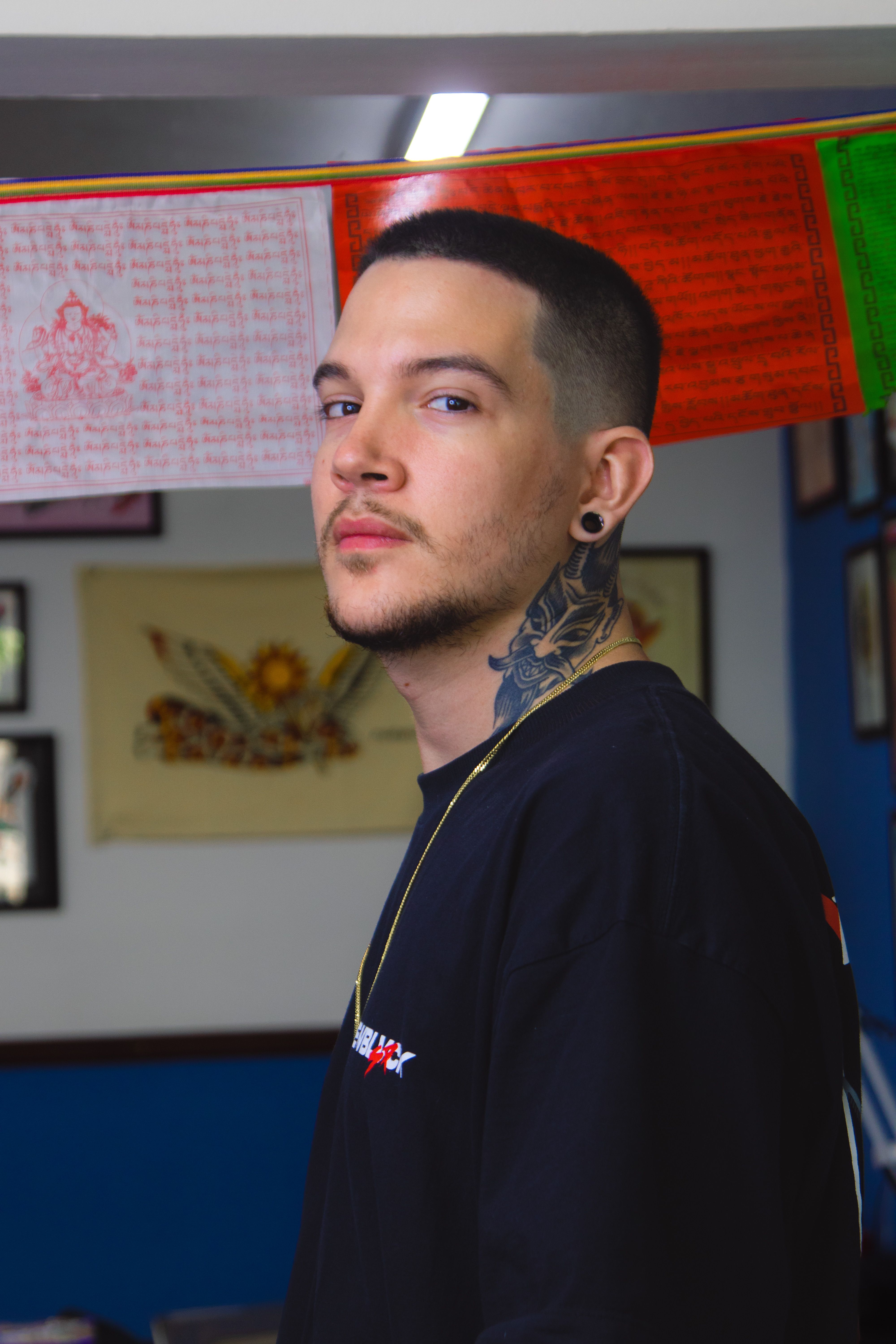 A man with a stylish haircut, tattoos, and mustache posing indoors with colorful flags in the background.
