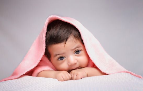 Charming portrait of a baby lying under a cozy pink blanket with a gentle gray background.
