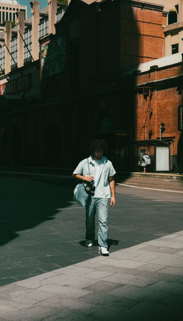 A person walks along a shadow-filled urban street, holding a camera, in front of Market City.