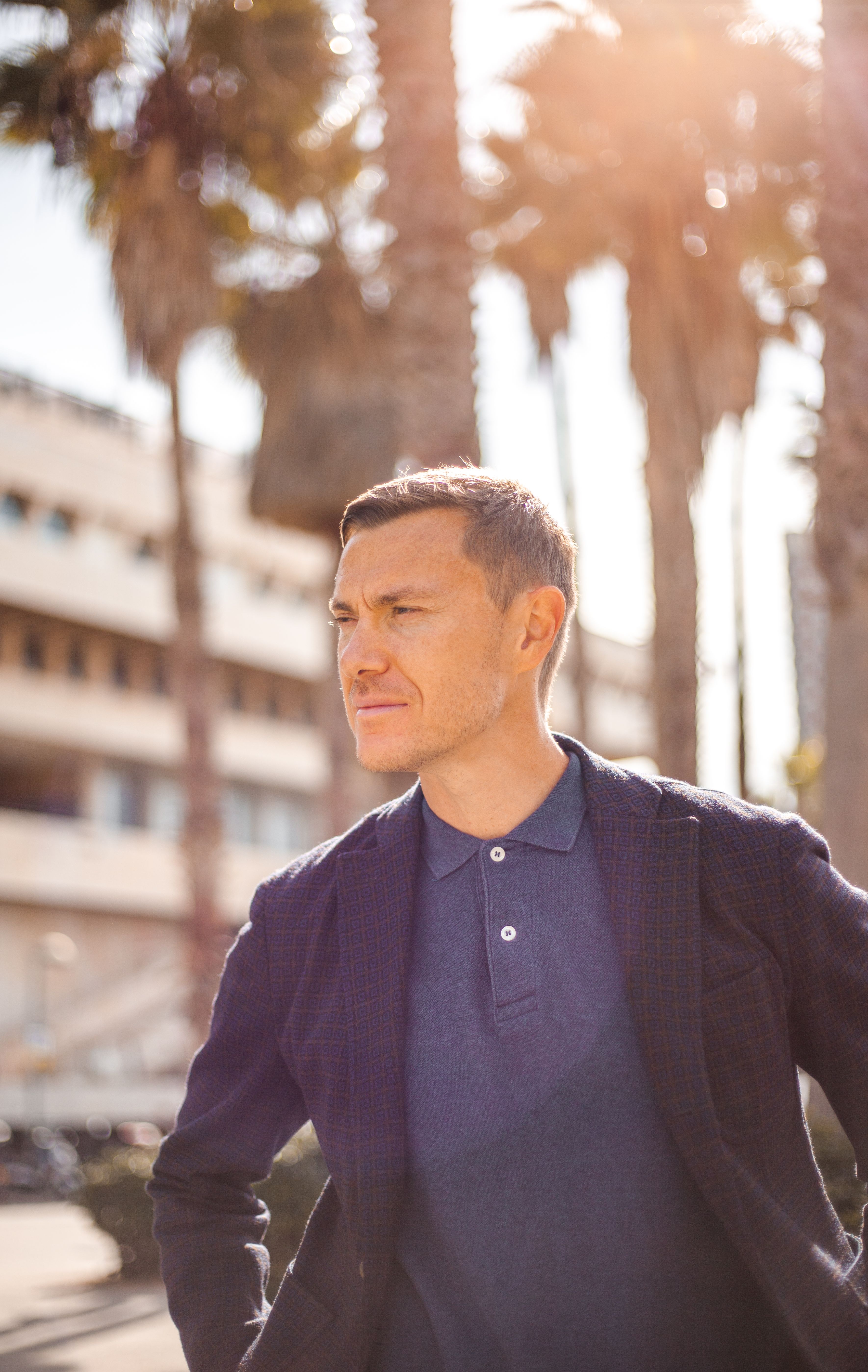 Portrait of a man in a stylish jacket under palm trees in Tel Aviv's warm sunlight.
