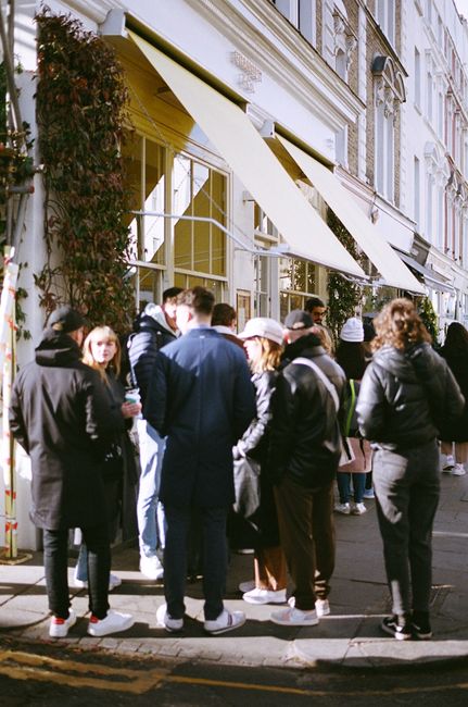 A diverse group of people gathered on a sunny city sidewalk near a cafe with yellow awnings.