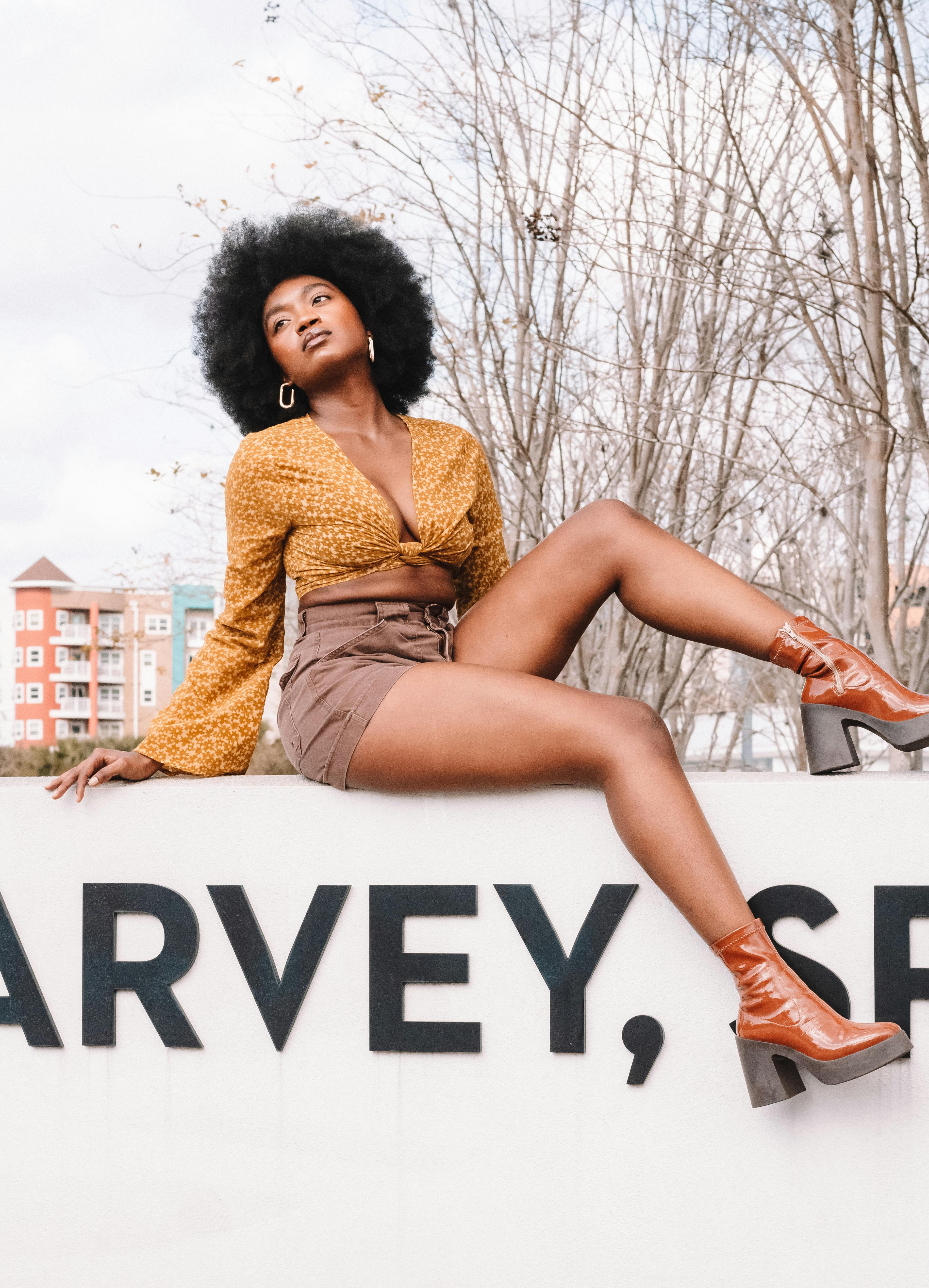 Young woman with afro hairstyle poses stylishly on a wall in autumn setting, showcasing fashion boots.