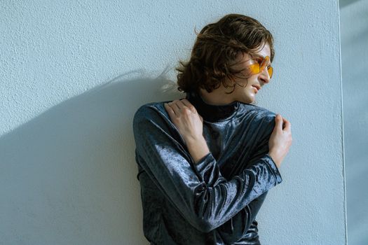 Fashionable man in a velvet blouse and yellow sunglasses posing against a white wall.