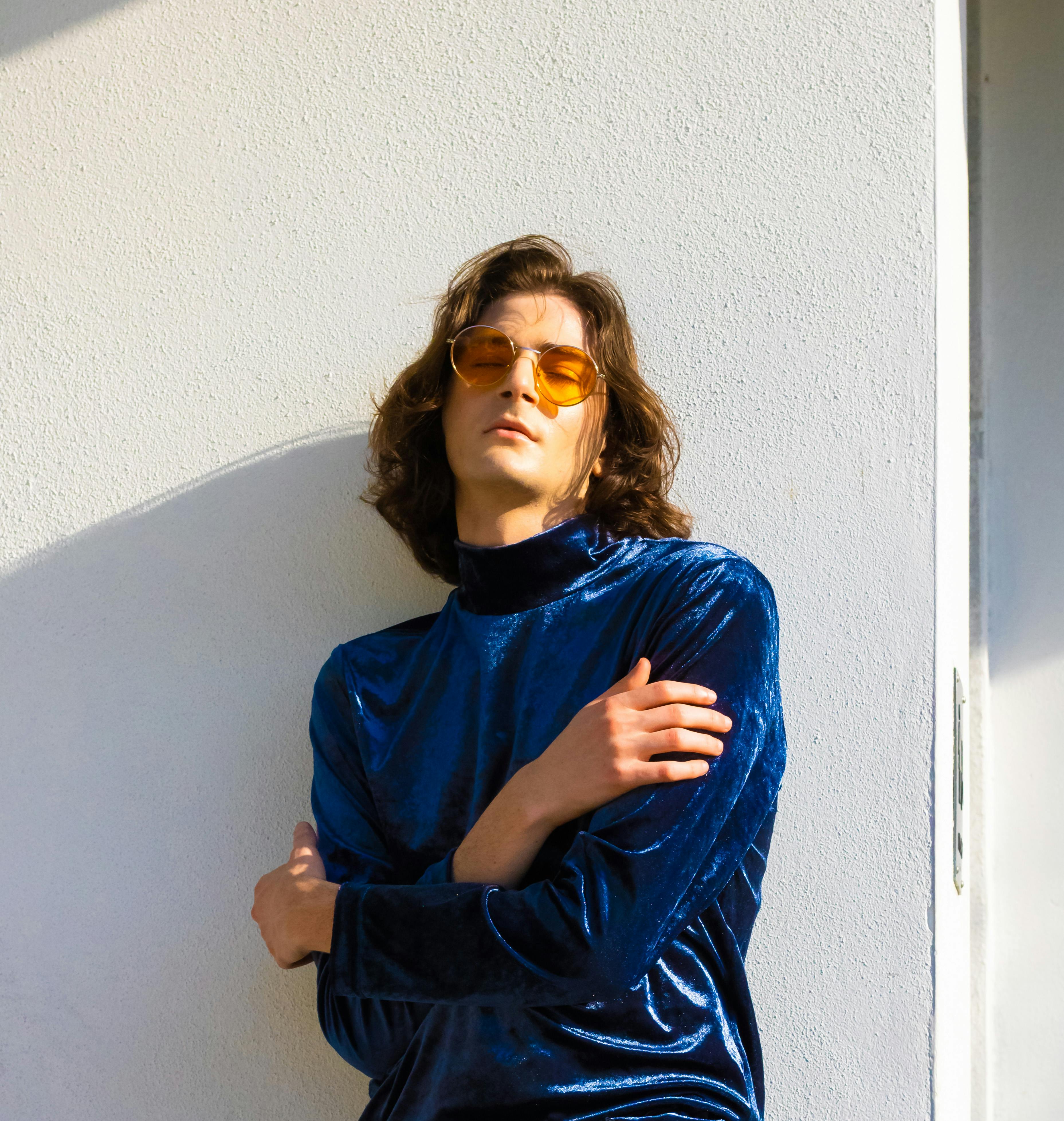 Fashionable young man in blue velvet top and orange sunglasses striking a pose against a sunlit wall.