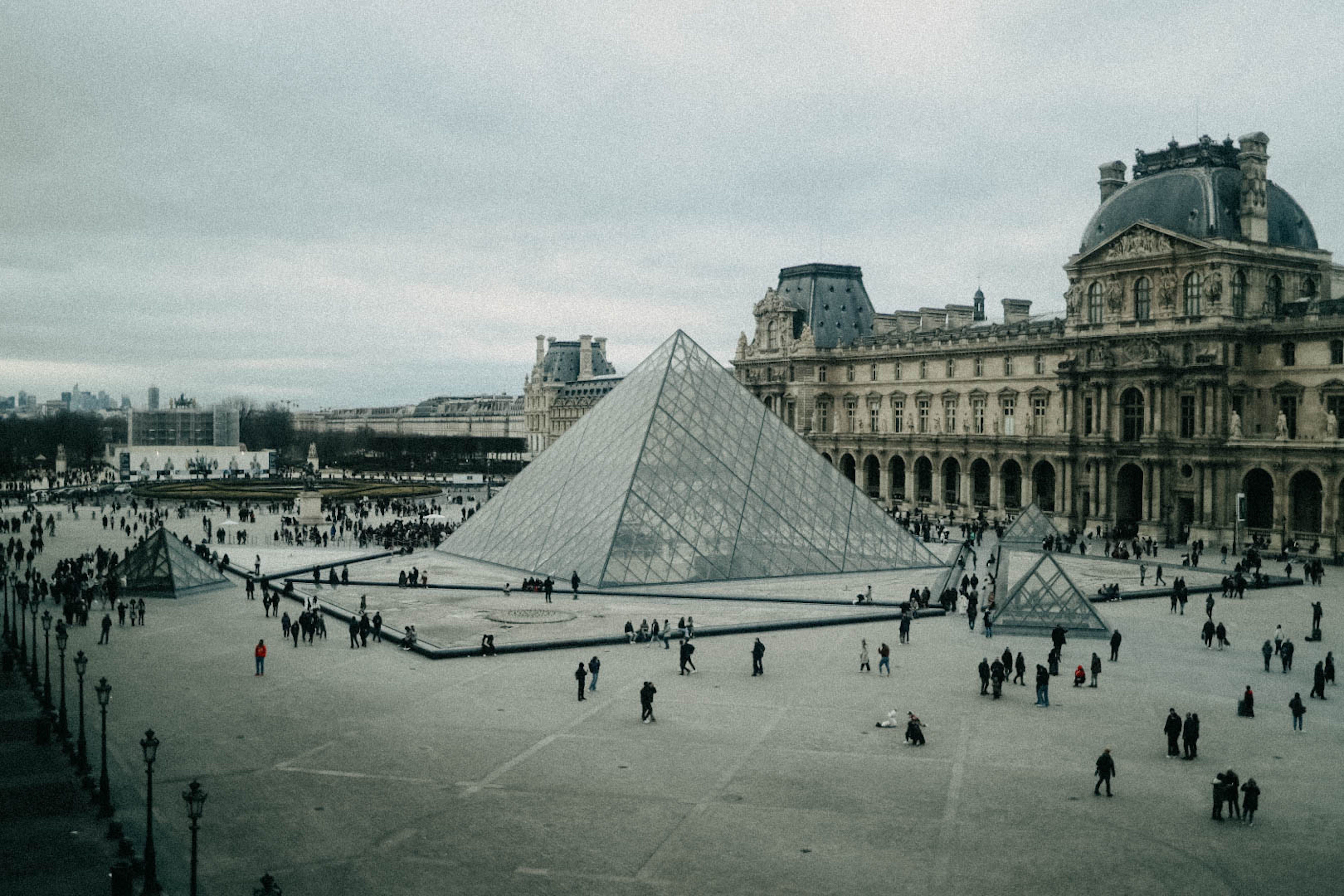Louvre Museum in Paris, France, with its glass pyramid, historic palace, crowds, and courtyard beneath soft overcast light