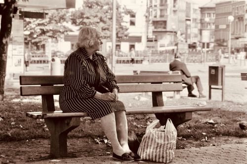 Free Sepia photo of an elderly woman sitting on a park bench with a bag, capturing a nostalgic urban scene. Stock Photo
