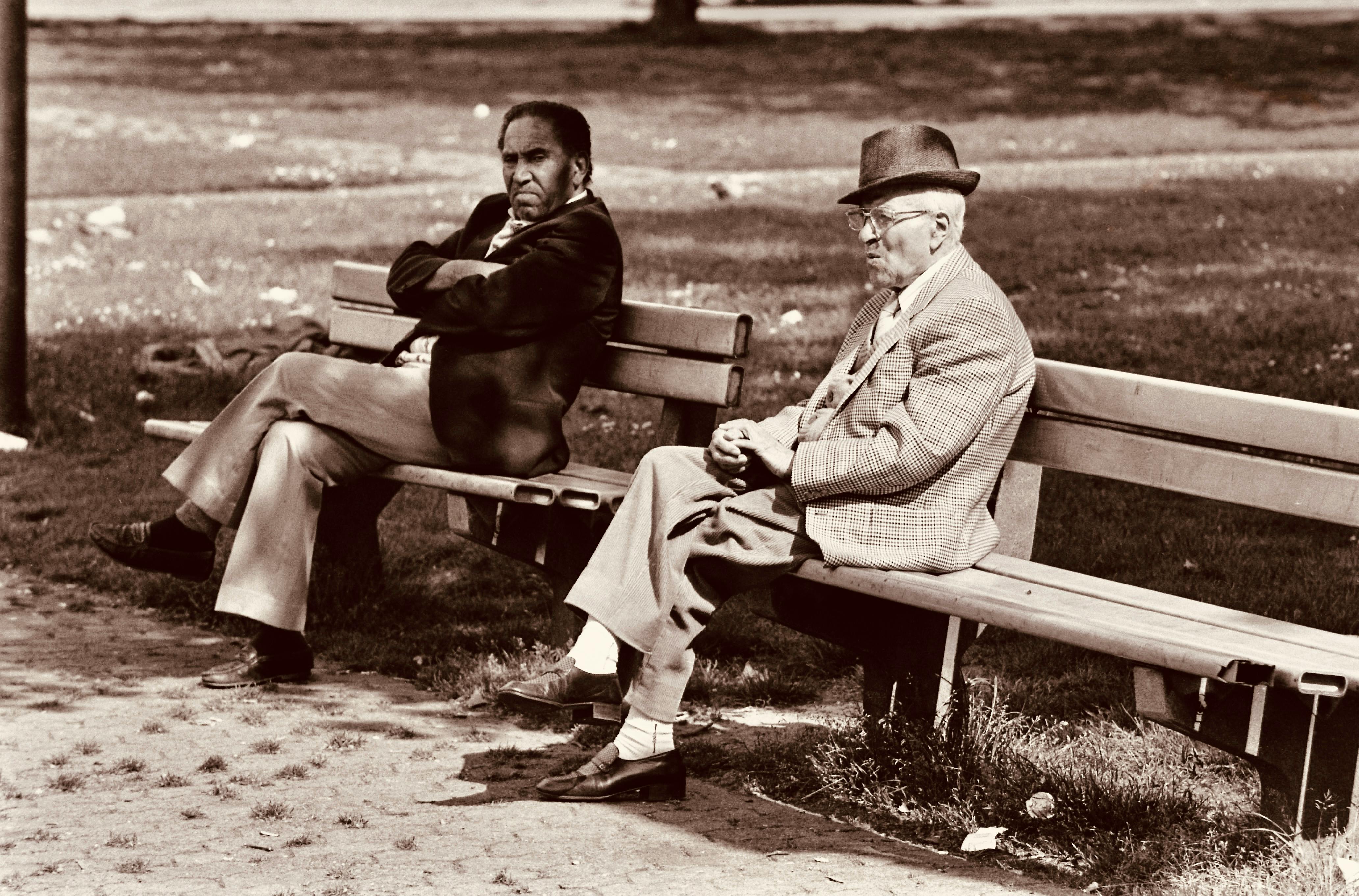 Two senior men sitting on benches in a park, chatting in vintage attire.