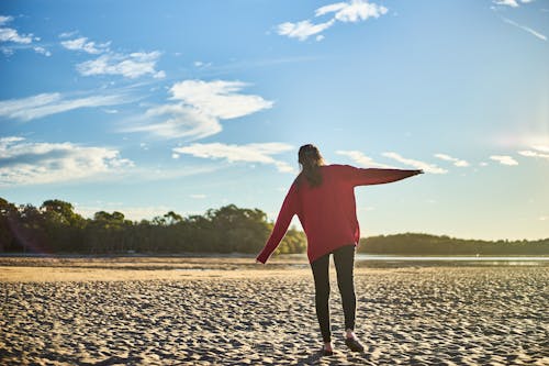Free Woman in Red Sweater and Black Pants on White Sand Near Body of Water During Daytime Stock Photo