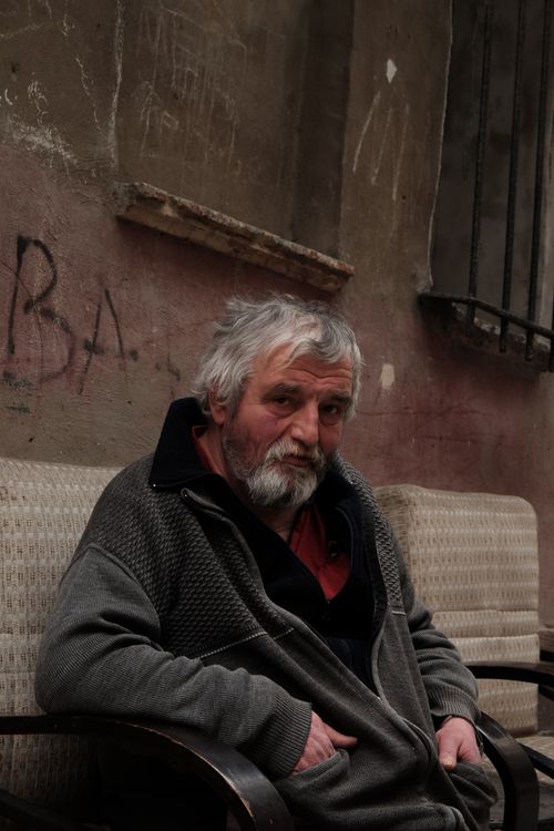 Free Elderly man with beard and mustache sitting outdoors on armchair in urban alley. Stock Photo