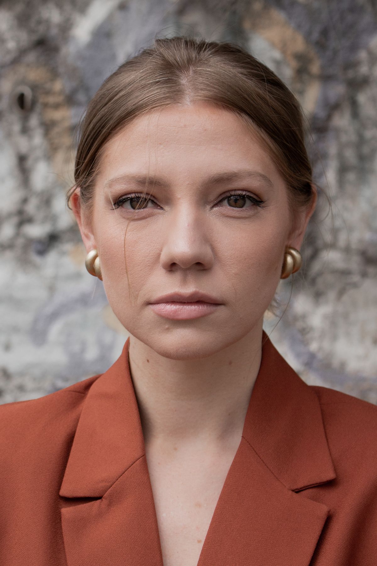Woman in a rust blazer wearing rounded gold earrings against a stone wall