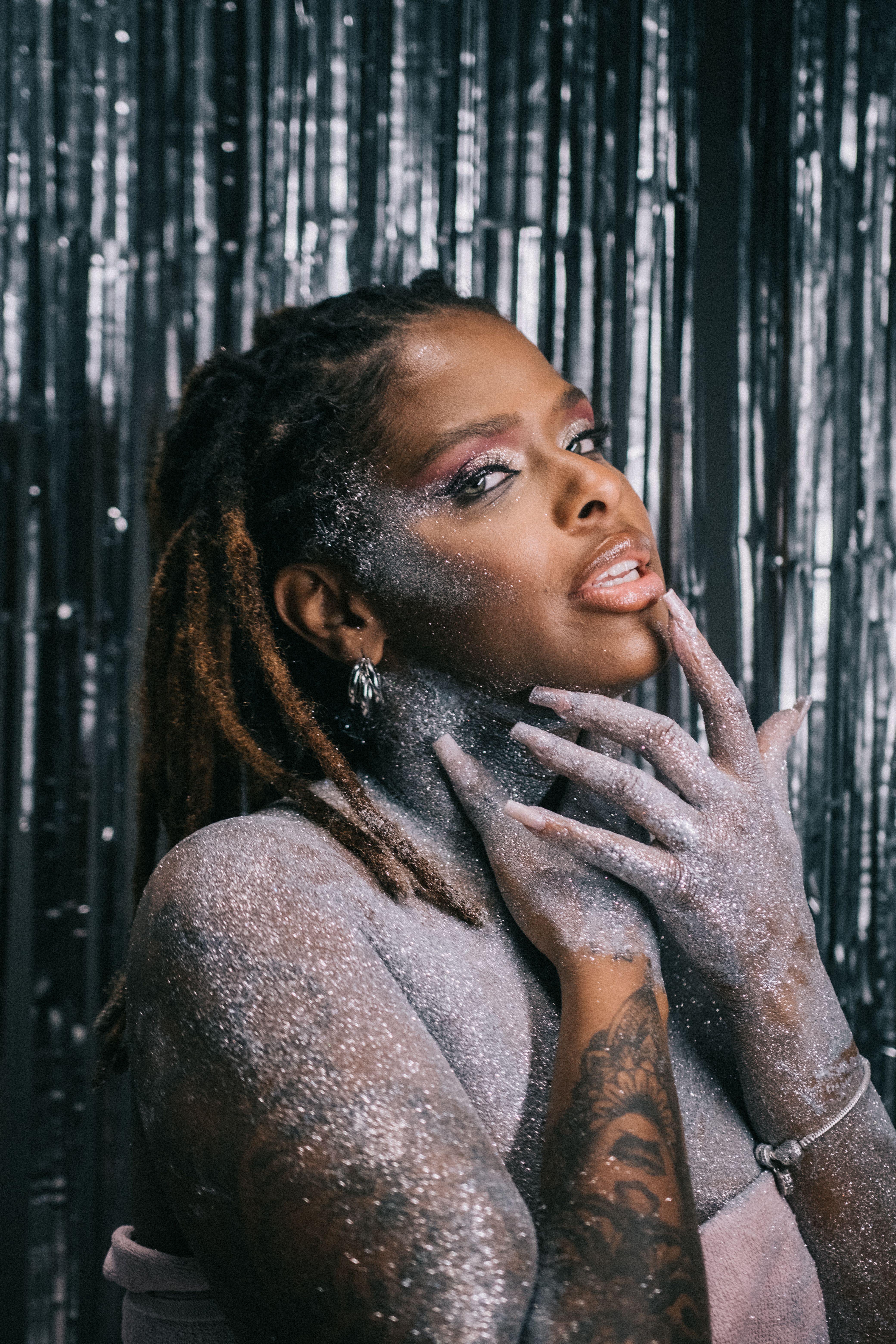 Artistic portrait of a glamorous woman covered in glitter against a silver curtain backdrop.