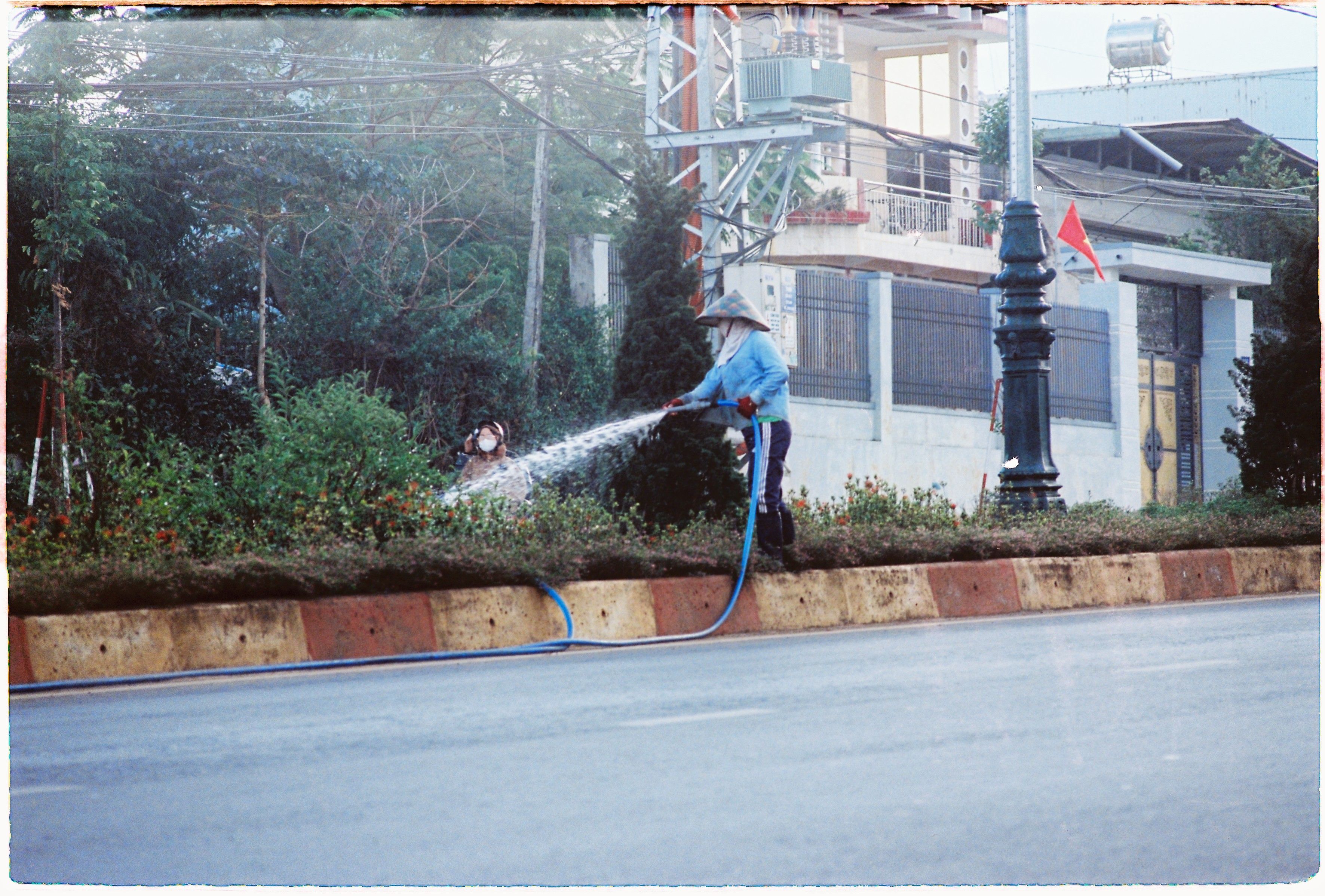 An urban gardener waters roadside plants in Pleiku, Vietnam, wearing a conical hat.