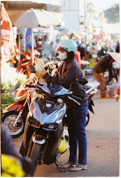 A woman wearing a helmet and mask stands by her scooter at a vibrant Vietnamese street market.
