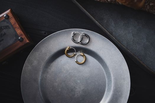 Stylish silver and gold hoop earrings arranged on a rustic metallic plate against a dark background.