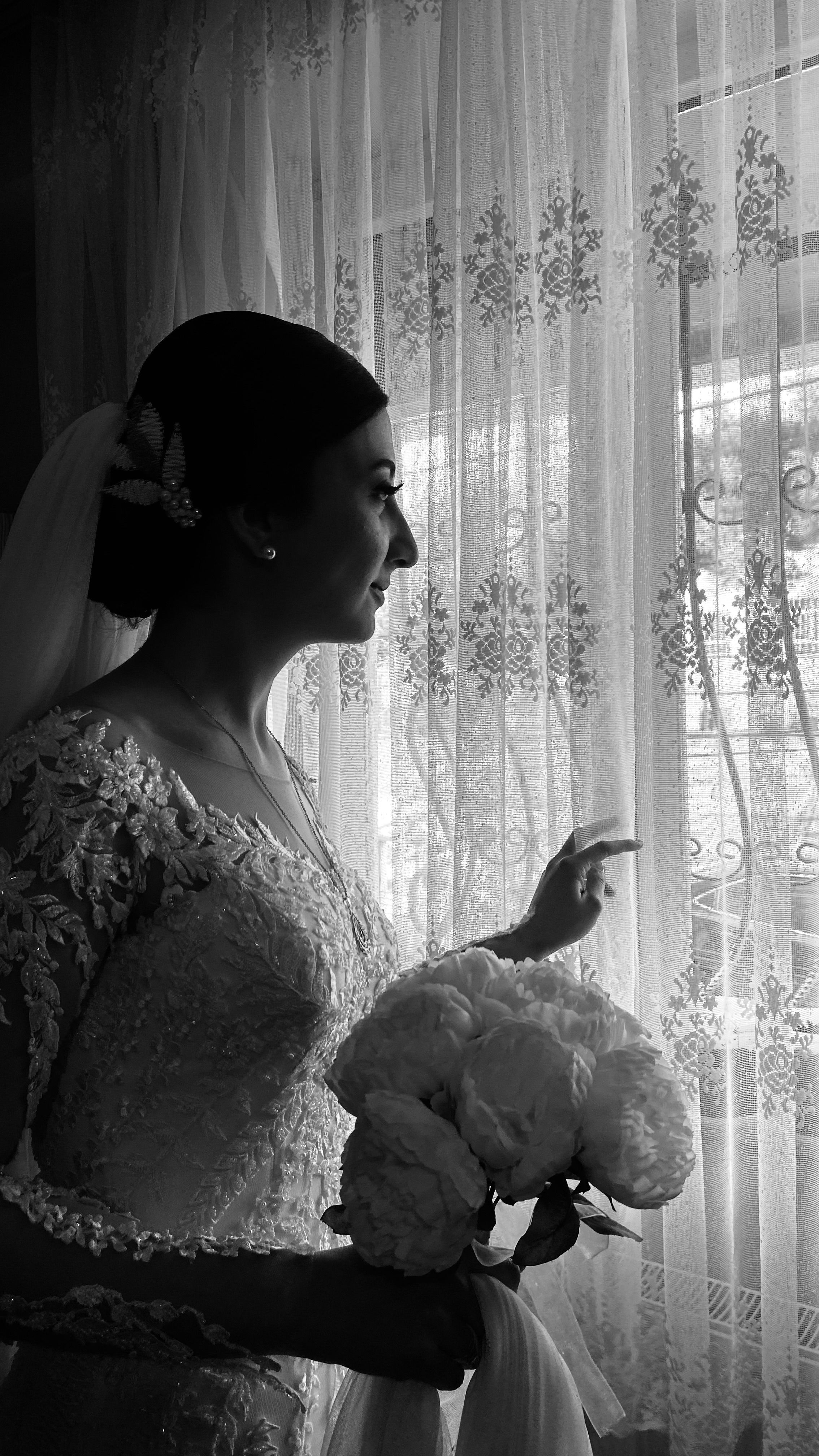 Black and white portrait of a bride in a lace gown holding flowers, looking out a window.
