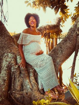 A joyful, pregnant woman wearing a crochet dress, sitting on a tree in a sunlit garden setting.