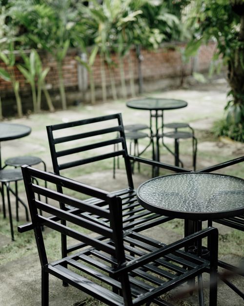 Free Serene outdoor café setting with empty black metal chairs and glass tables amidst lush green plants. Stock Photo