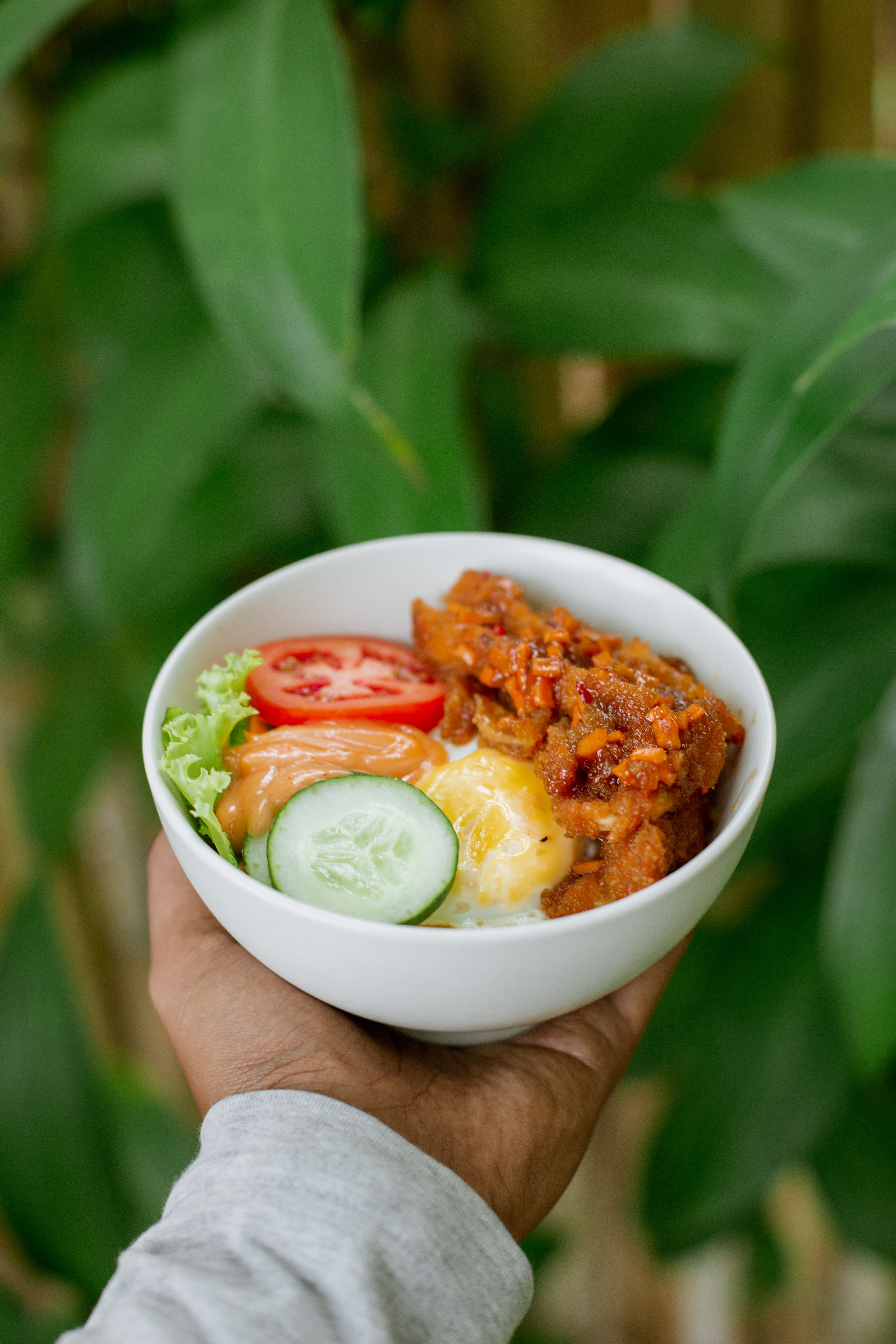 A fresh bowl with vegetables and protein, held outdoors surrounded by greenery.