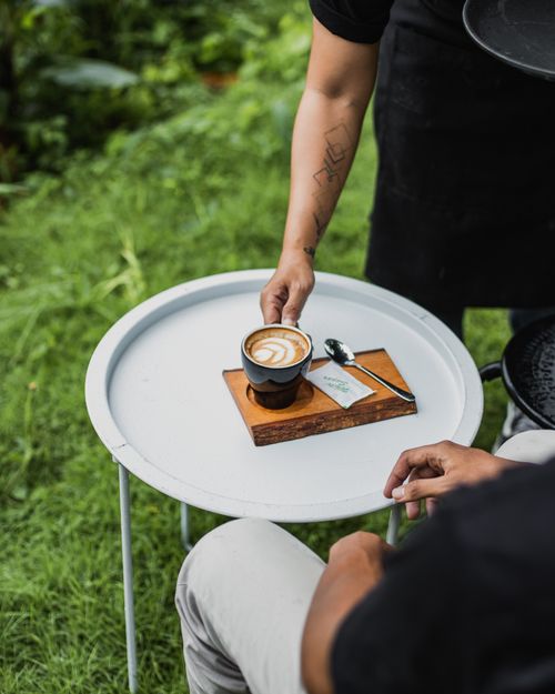 Free A barista serving a cup of latte coffee with art on a small round table outdoors. Stock Photo