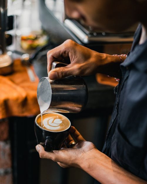 Free Close-up of a barista pouring milk into a cappuccino, creating latte art in a cozy café setting. Stock Photo