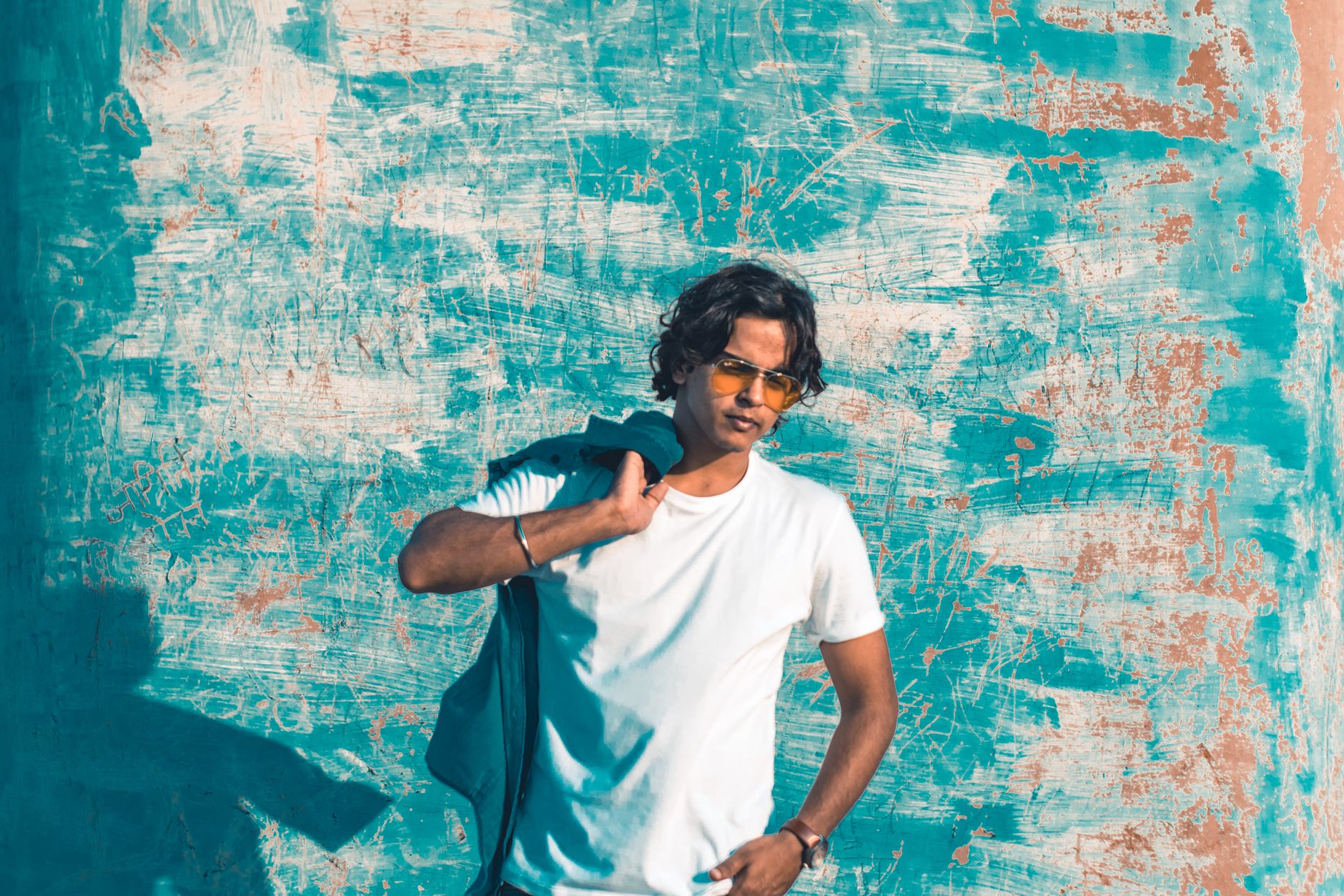 Young man in sunglasses leaning against a textured blue wall, exuding cool style.