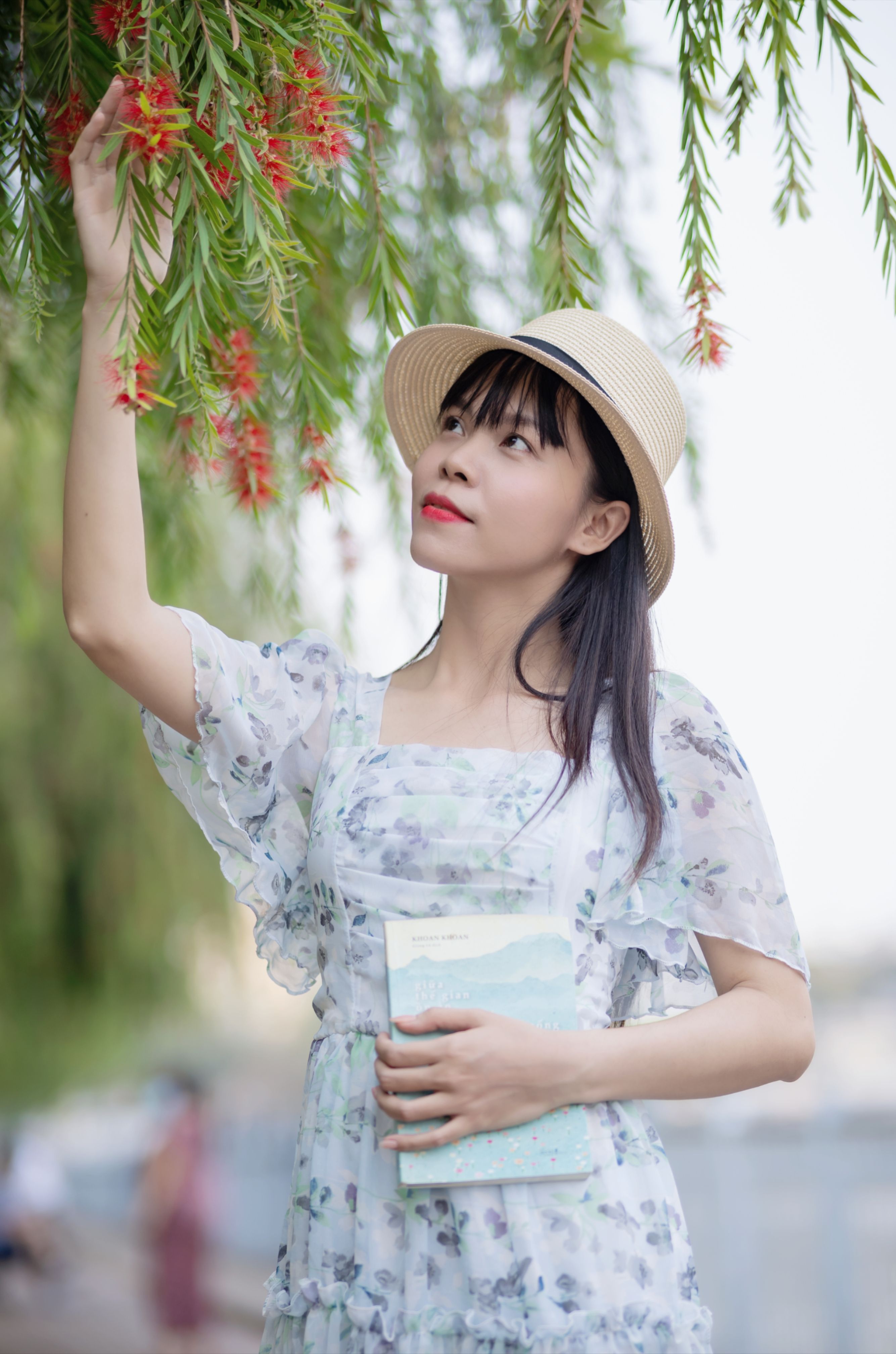 Woman in a floral dress and straw hat, holding a book, touching blossoms on an outdoor tree.