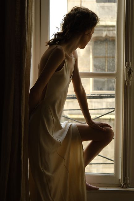 A young woman in a dress sits by a window, gazing outside thoughtfully.