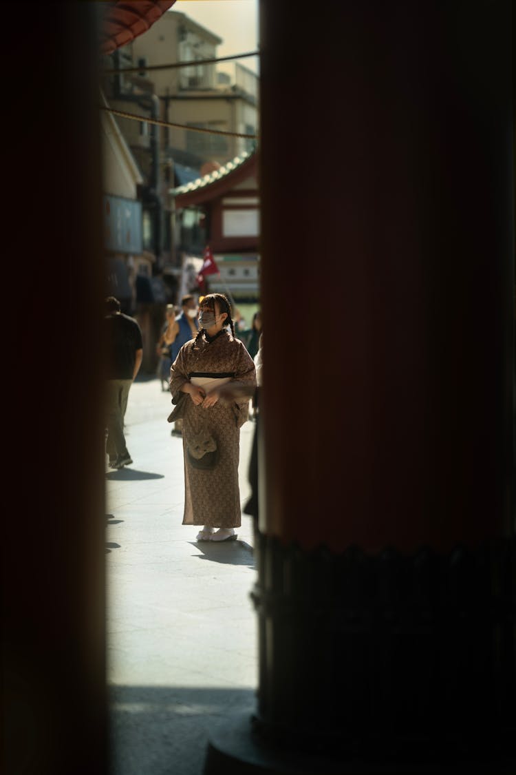 Candid Photo Of A Woman In A Traditional Gown And Face Mask Standing On A Street 