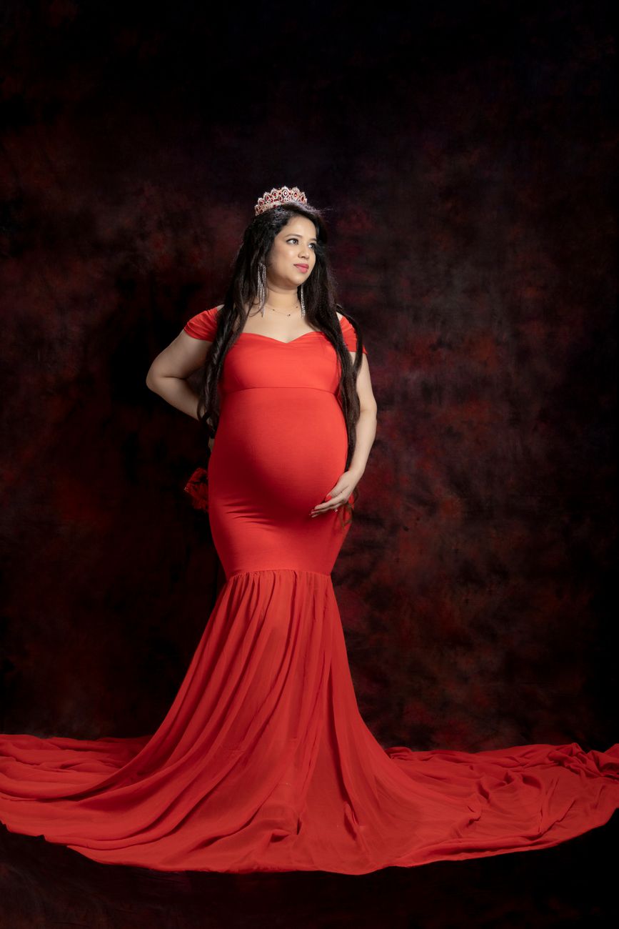 A glamorous pregnant woman in a long red dress posing elegantly in studio setting.