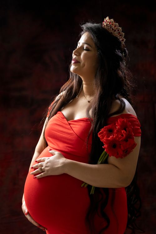 Free A pregnant woman in a red dress with a crown, smiling and holding red flowers in a studio shoot. Stock Photo