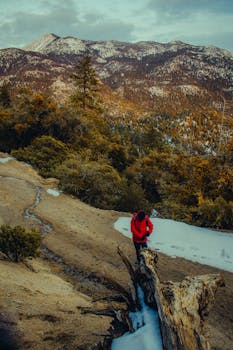 A person in a red jacket hikes along a scenic mountain trail with snow patches and a forested landscape.