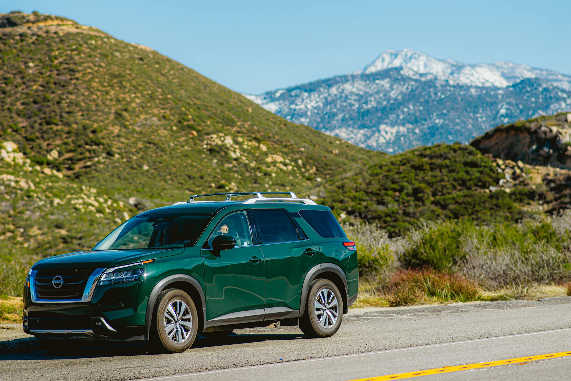 Green Nissan Pathfinder SUV on a road near lush mountains against a clear blue sky.