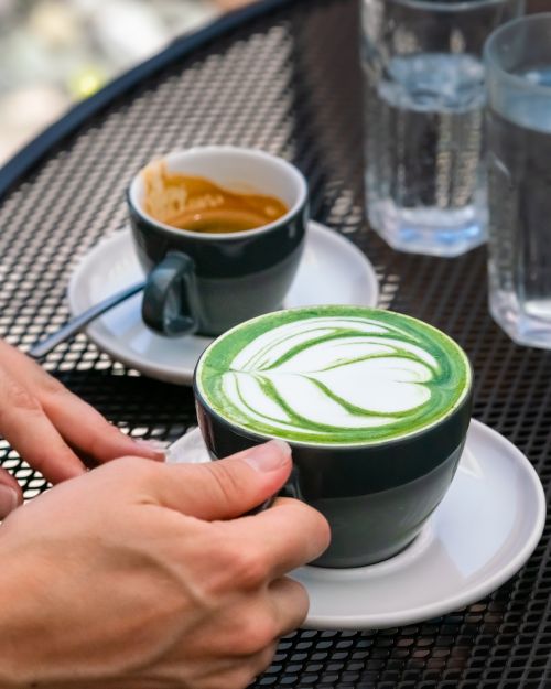 Free A person's hands holding a matcha latte with milk foam art, next to an espresso in a café setting. Stock Photo