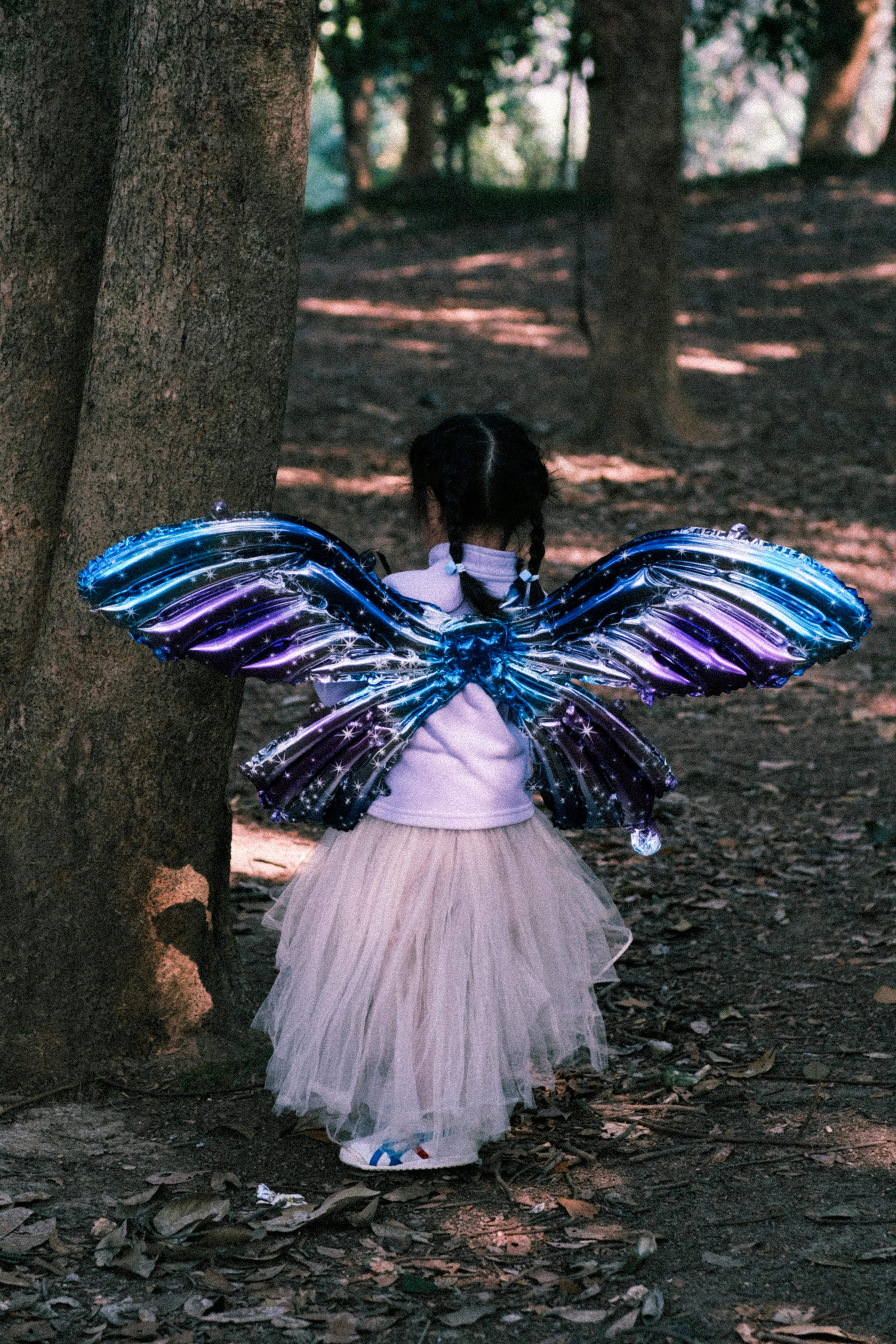 A young girl in a fairy costume with wings in a wooded park, seen from behind.