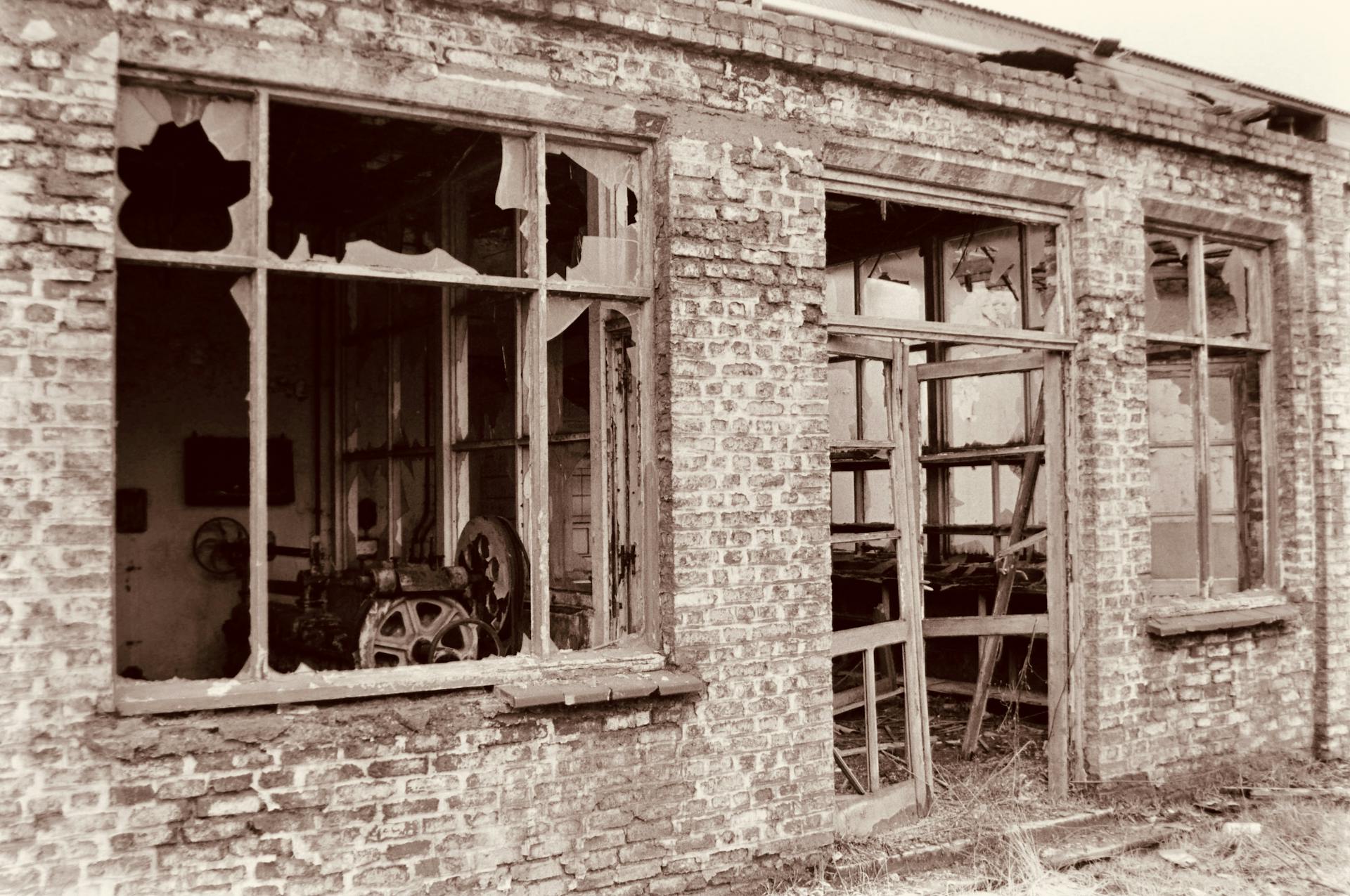 A derelict brick building with broken windows and rusting machinery inside, in sepia tone.