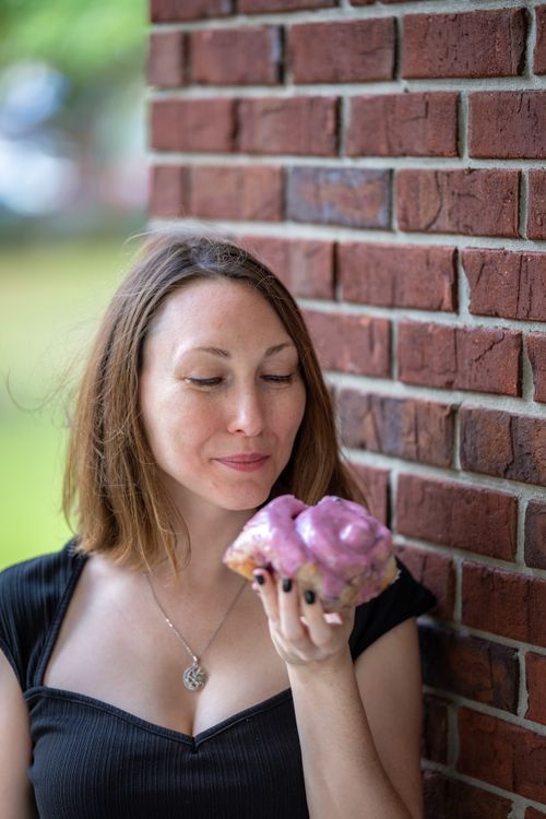 Free A woman holding a pink frosted donut while standing next to a brick wall outdoors. Stock Photo