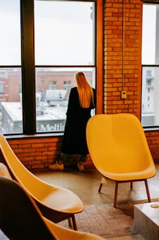 A woman in a black coat stands by a large office window overlooking an urban setting.