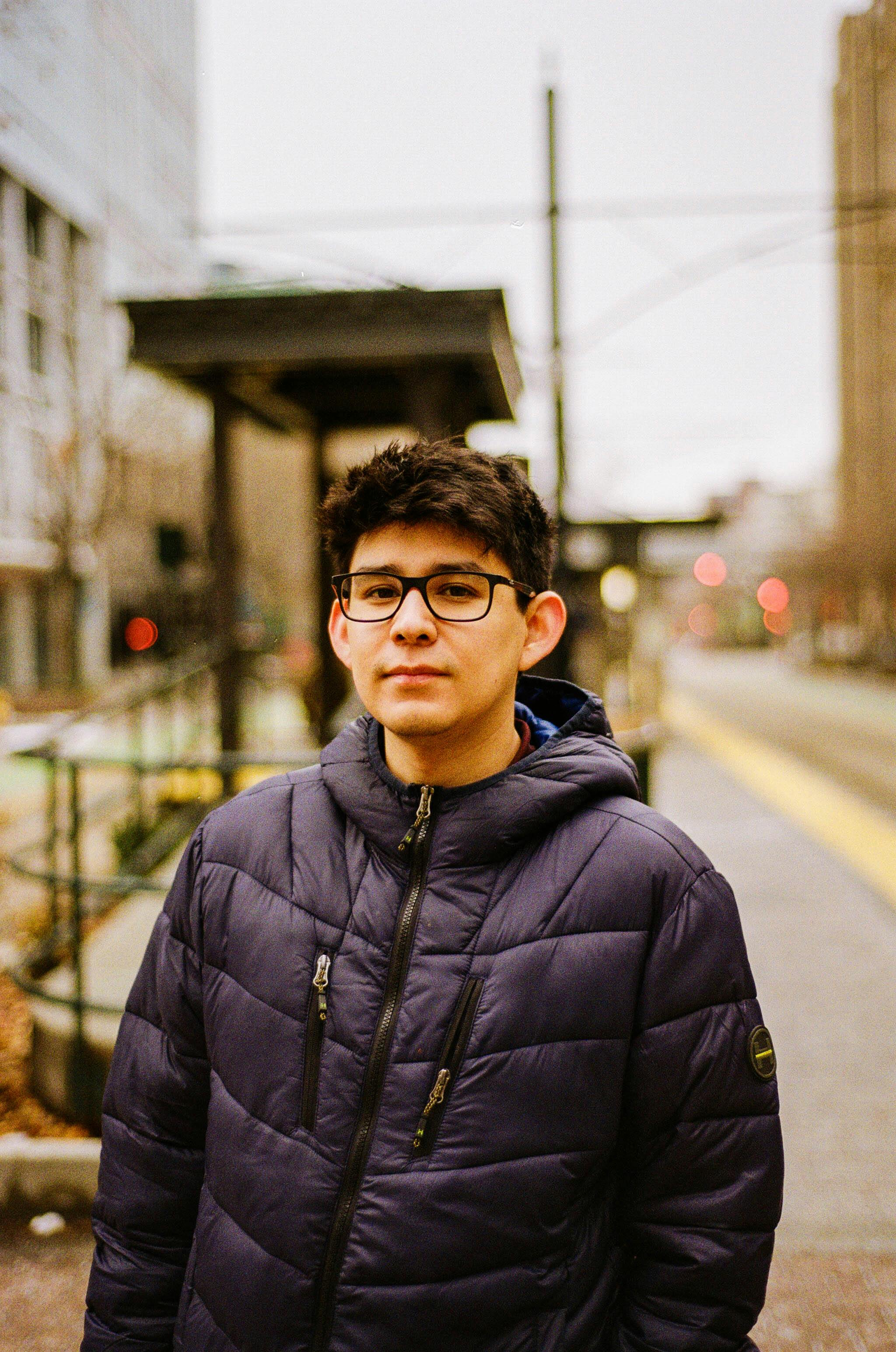 Young man with eyeglasses in a jacket outdoors in an urban environment.