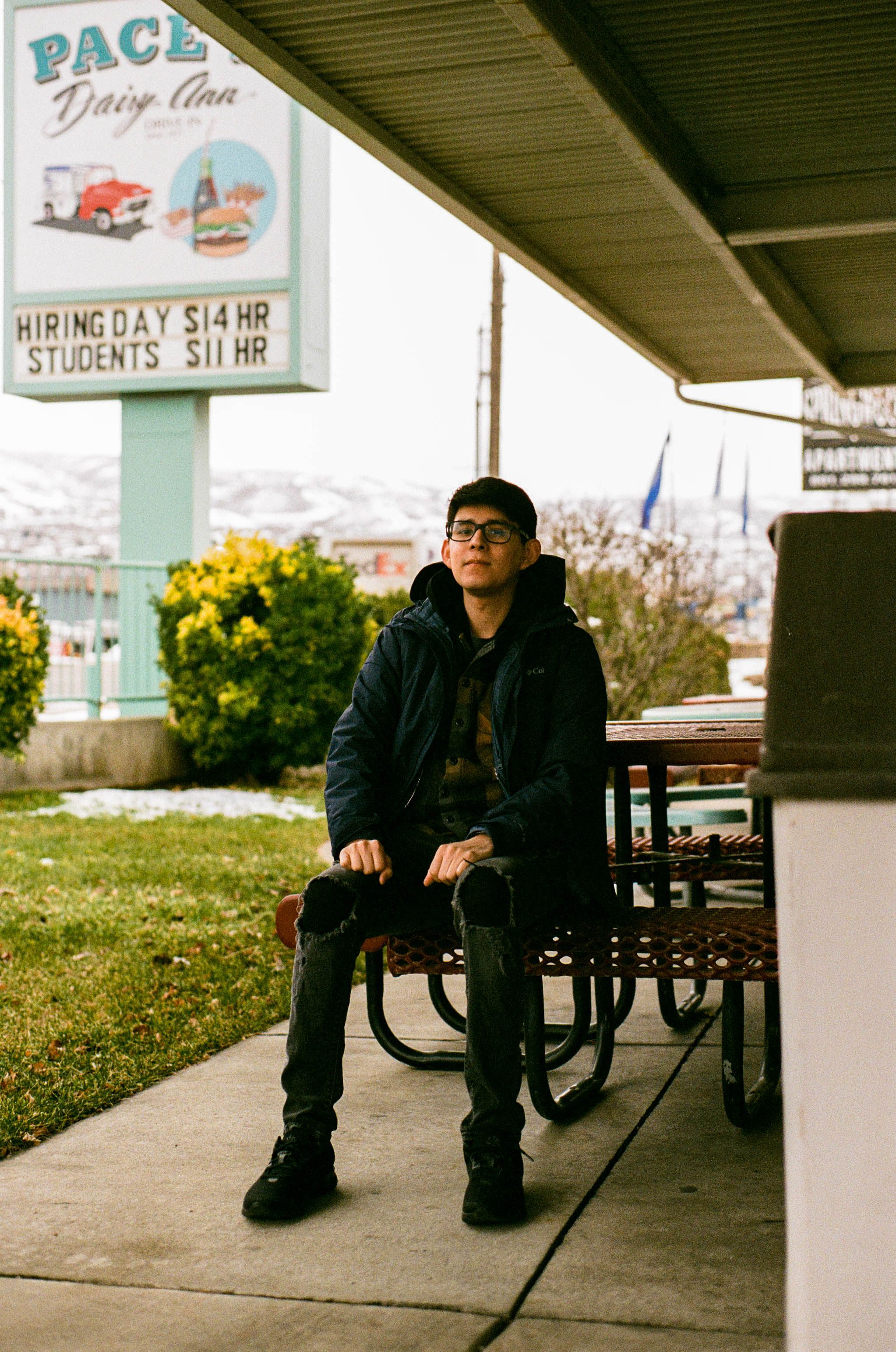 A young man sitting at an outdoor diner, looking relaxed with snow-covered mountains in the background.