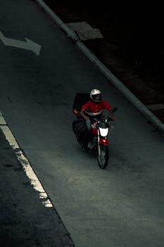 Aerial view of a delivery rider in a helmet on a red motorcycle navigating an urban road.