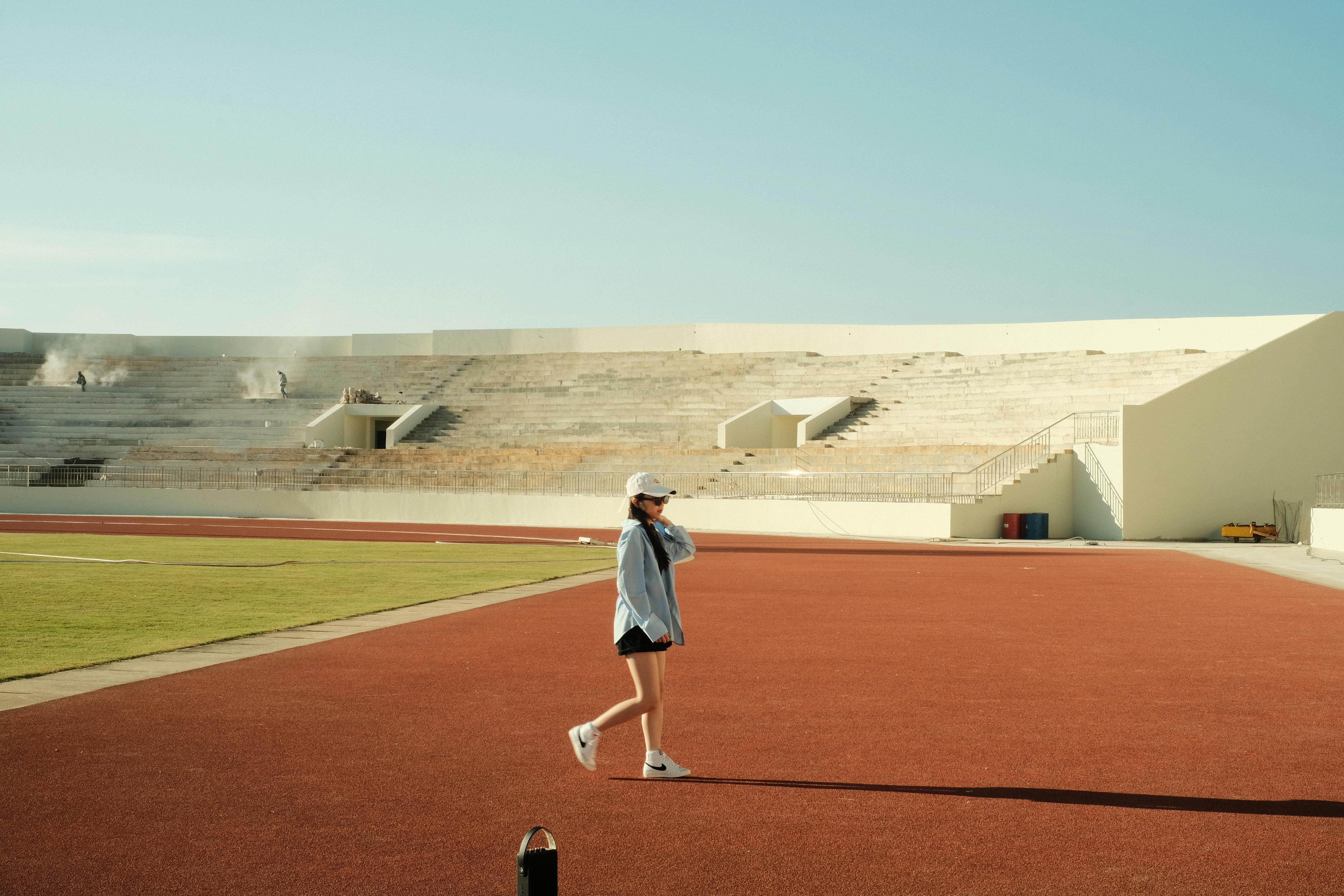 A young woman in casual attire walks across an empty stadium track on a sunny day.