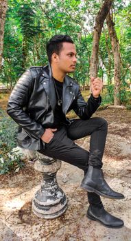 Man in a leather jacket casually sitting on a stone pillar in a forest setting.